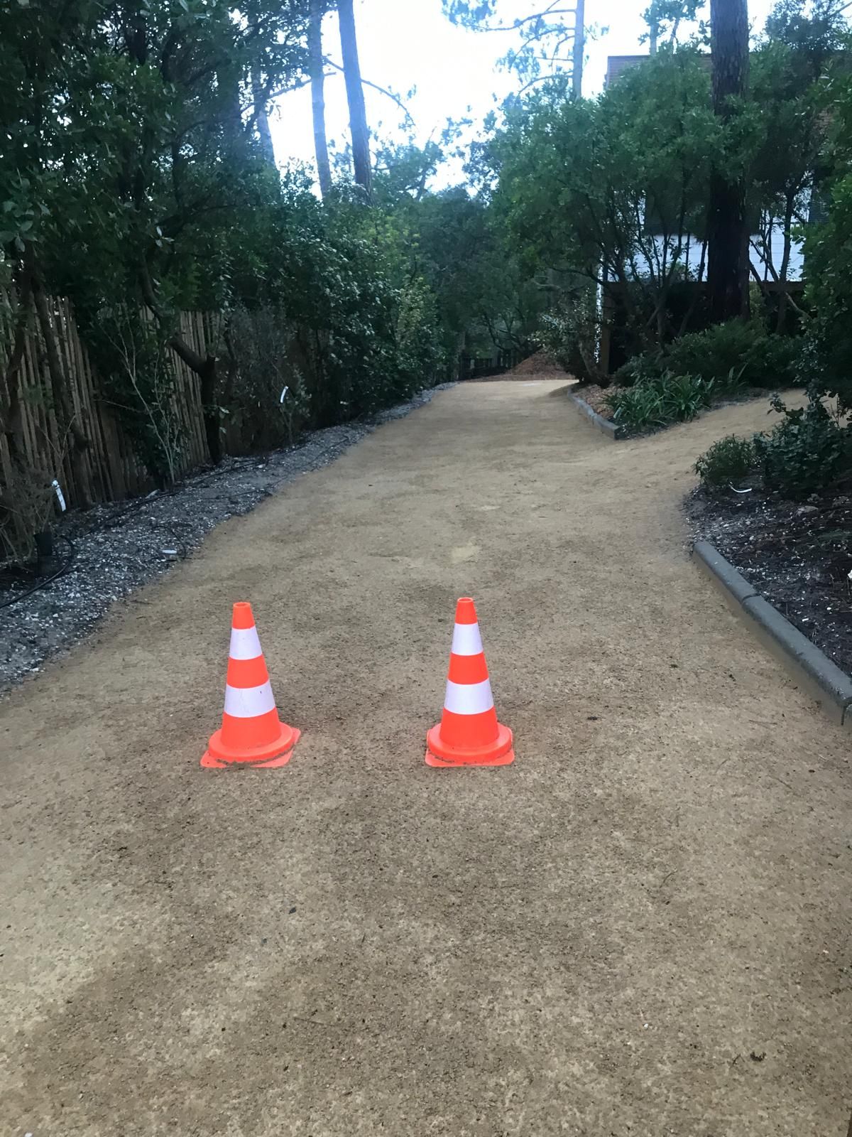 Deux cônes de signalisation orange et blanc bloquent un chemin de terre fraîchement stabilisé bordé d'arbres et d'une palissade en bois.