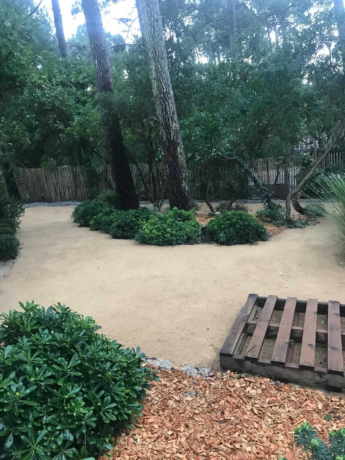 Allée de jardin en sable ocre, bordée de haies vertes et d'arbres, avec une clôture en bois et une palette sombre au premier plan.