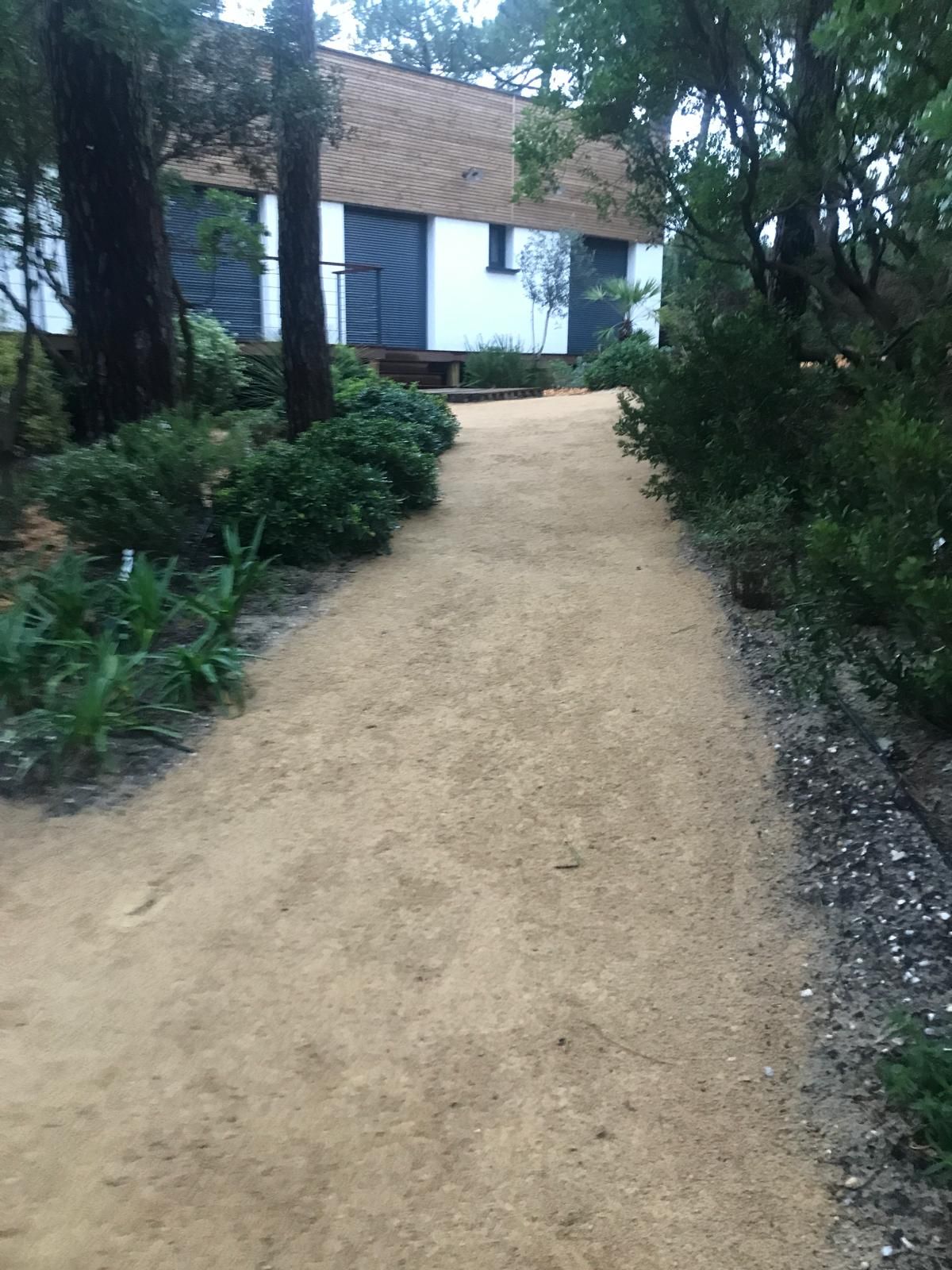 Allée de gravier ocre menant à une maison moderne avec façade blanche et bardage bois, entourée d'arbres et de végétation dense.