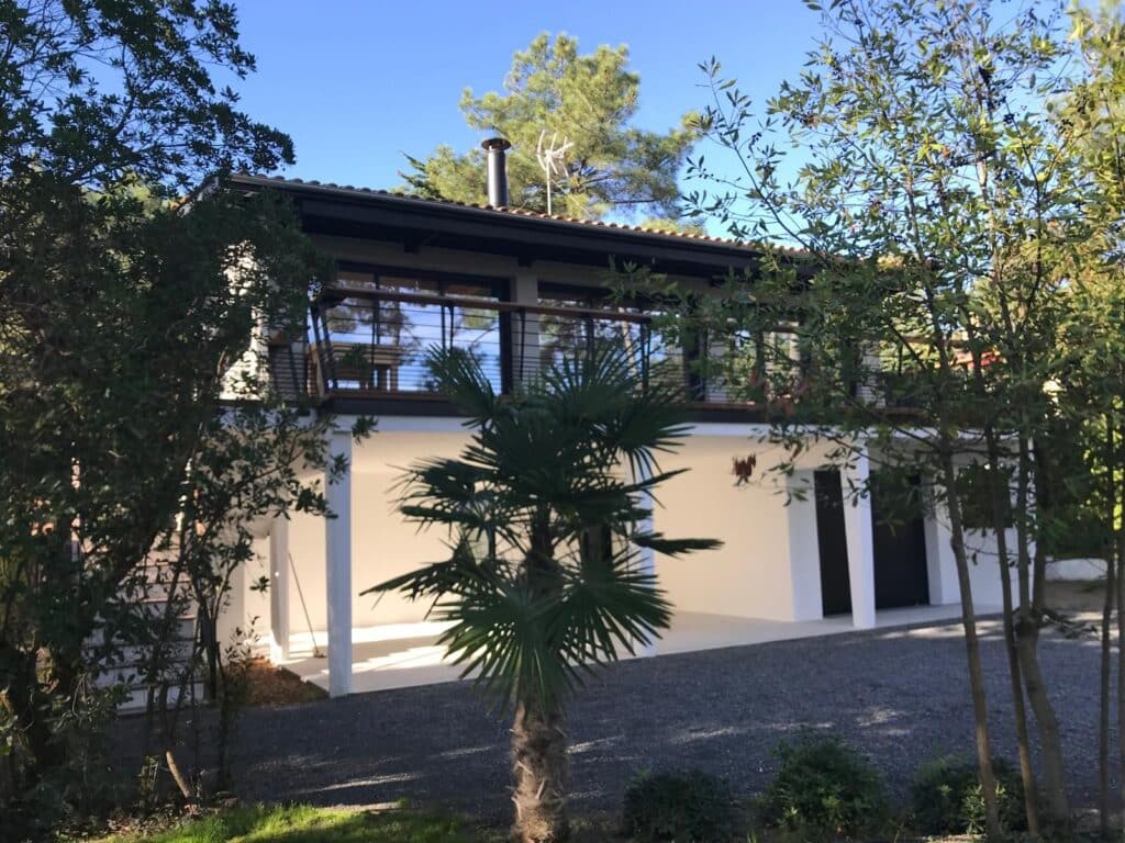 Maison Moderne Bois et Verre au Milieu de la Nature Maison moderne blanche à deux étages avec balcon et grandes baies vitrées, entourée d'arbres verts et d'un petit palmier au premier plan sous un ciel bleu.