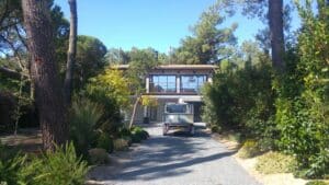 Maison moderne en lisière de forêt, accès par allée gravillonnée, une remorque de chantier est garée devant le garage.