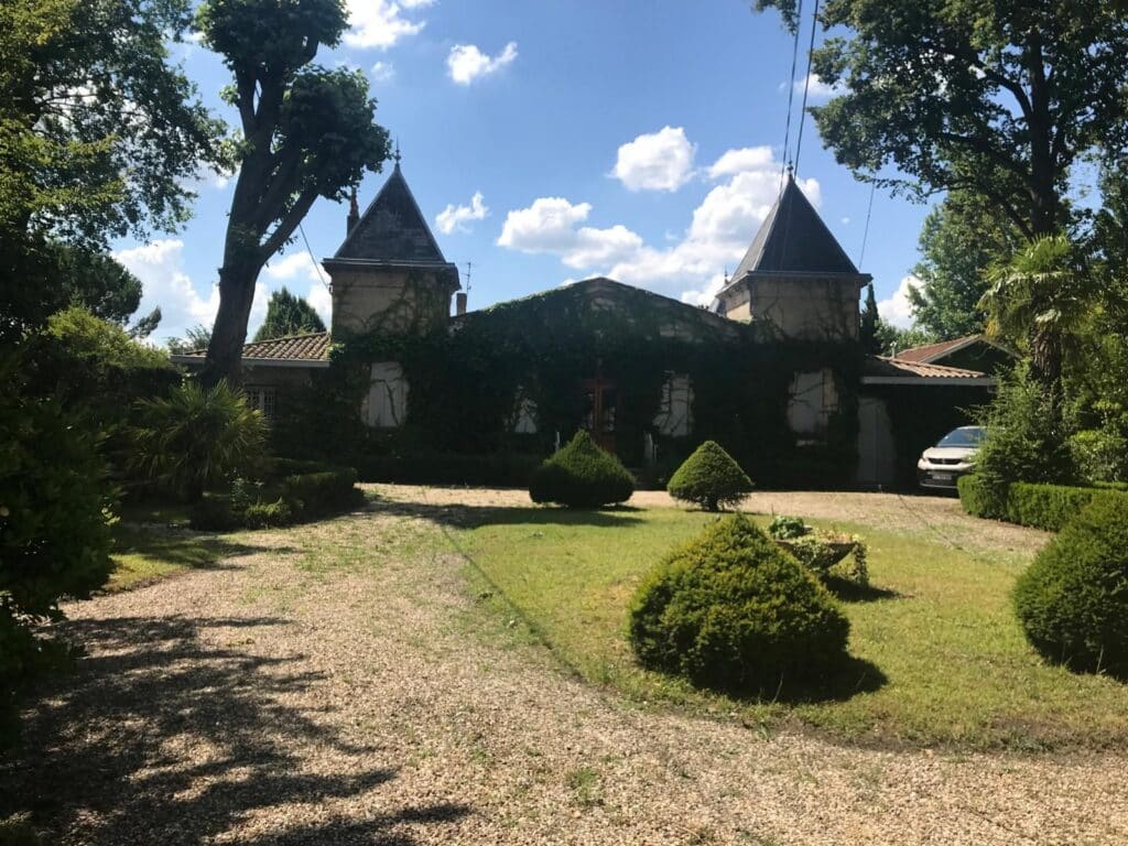 Maison Ancienne avec Tourelles et Jardin luxuriant Façade d'une maison ancienne en pierre recouverte de lierre, flanquée de deux tourelles coniques, avec un jardin et une allée en gravier par temps ensoleillé.